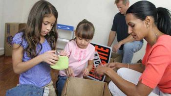 Two kids help their mom unpack a box after a move. Their dad does the same in the background.